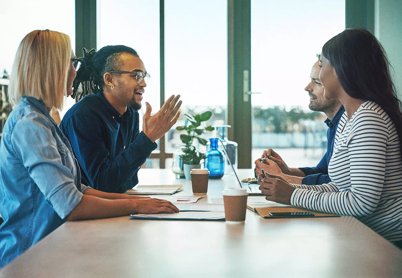 A group of professionals engaged in a discussion around a table in a bright, modern office space. The setting includes large windows with natural light, a glass water bottle, and disposable coffee cups. The atmosphere suggests collaboration and teamwork.